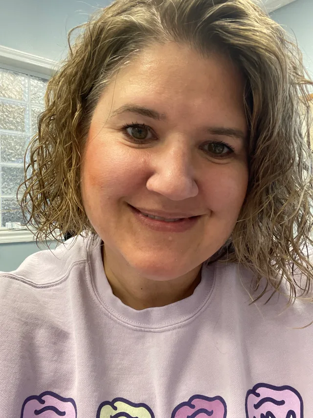 Woman smiling in a selfie style photo with a purple sweatshirt and blonde, curly hair